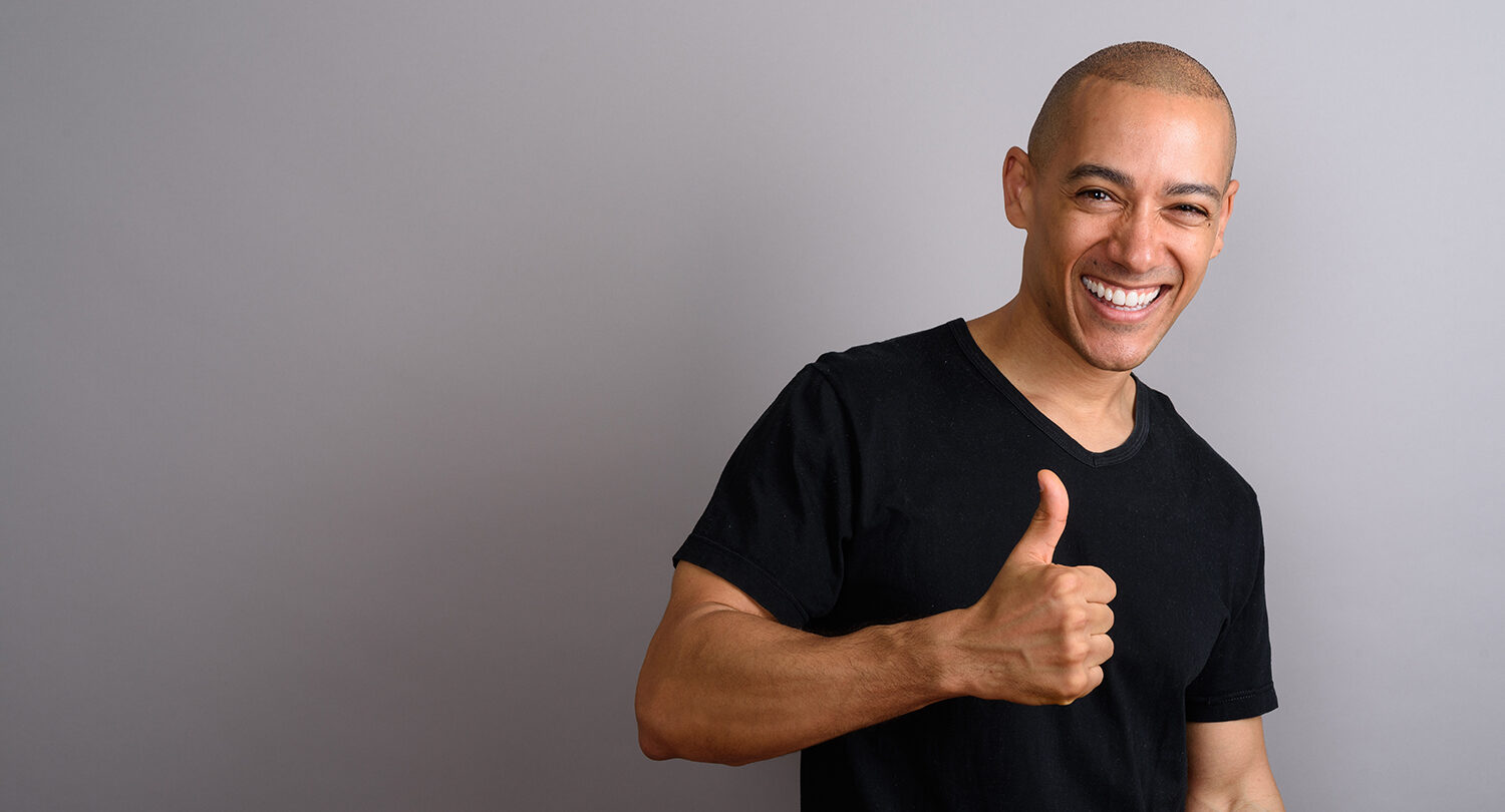 Studio shot of handsome bald man wearing black shirt against gray background holding thumb up